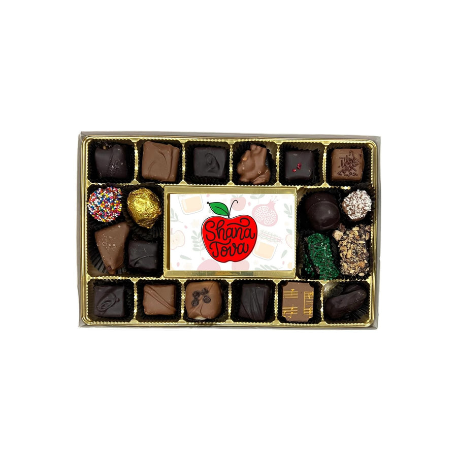 A boxed assortment of chocolates and truffles arranged in a gold tray, with a central white card featuring a red apple logo that reads 'Shana Tova'.